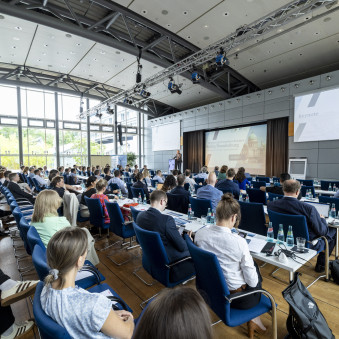 Großer Konferenzsaal mit Bühne im Hintergrund. Im Vordergrund ist das Publikum zu sehen, das sitzend aufmerksam den Vorträgen folgt.