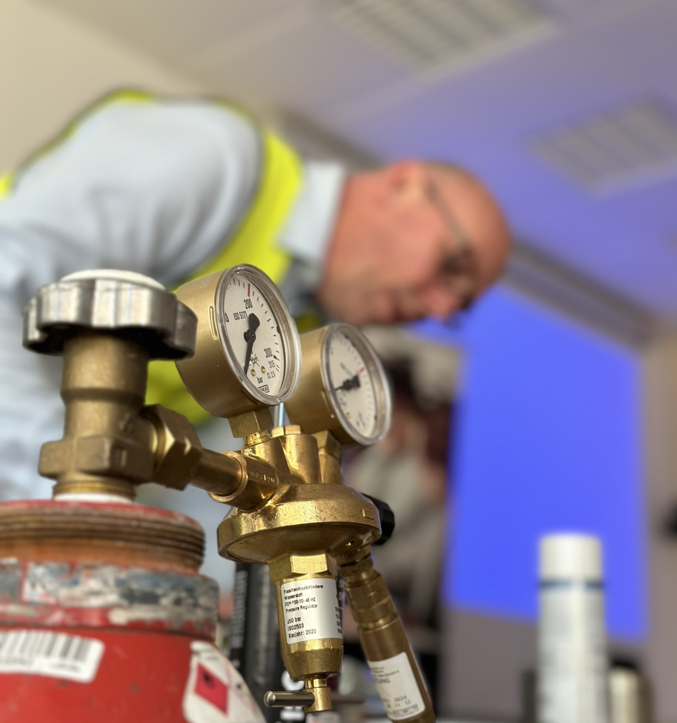 Druckminderer-Ventil an einer roten Gasflasche im Vordergrund, mit einer unscharfen Person in Sicherheitsweste im Hintergrund.